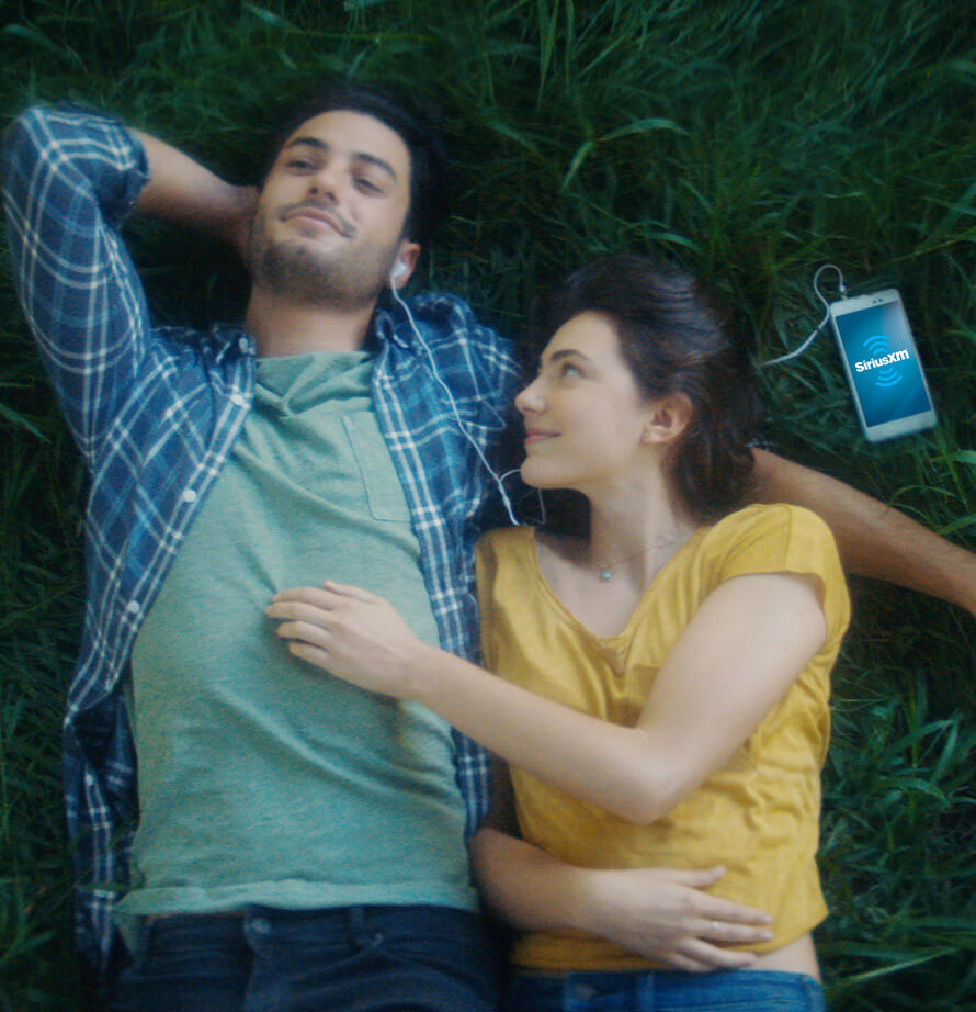 A man and woman lying on the grass sharing headphones while listening to SiriusXM.