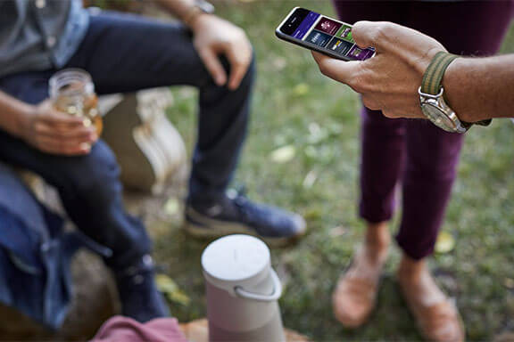 Un groupe de gens autour d’un haut-parleur Bose utilisant l’application SiriusXM démontrée sur un téléphone pour le contrôler.