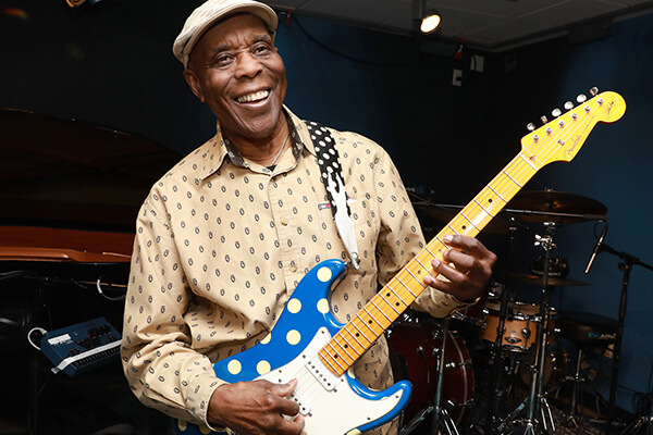 An image of Buddy Guy holding a blue Fender guitar with polkadots.