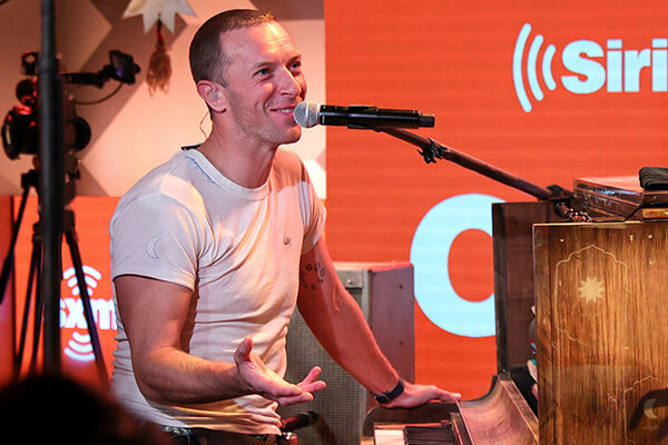 An image of Chris Martin of Coldplay sitting behind a piano smiling.