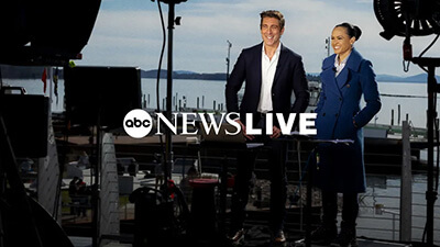 Two news anchors stand outdoors with waterfront and boats in the background, surrounded by studio lights and equipment. The ABC News Live logo and a What’s New banner are overlaid on the image, highlighting fresh updates from the scene.