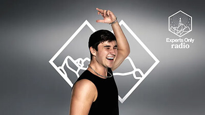 A young man in a sleeveless black shirt smiles and raises one arm in front of an abstract geometric background with "What’s New" and Experts Only radio text and logo on the right.