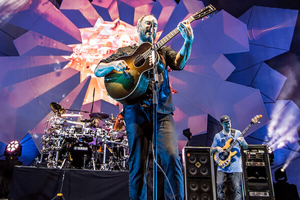 Un groupe se produit sur scène avec un éclairage dramatique et des formes géométriques en arrière-plan. Le chanteur joue de la guitare à l'avant, tandis qu'un batteur et un bassiste jouent derrière lui.