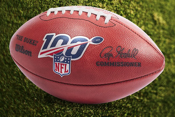 Gros plan d'un ballon de football officiel de la NFL portant le logo 100, la marque Wilson et la signature du commissaire, reposant sur une pelouse verte.