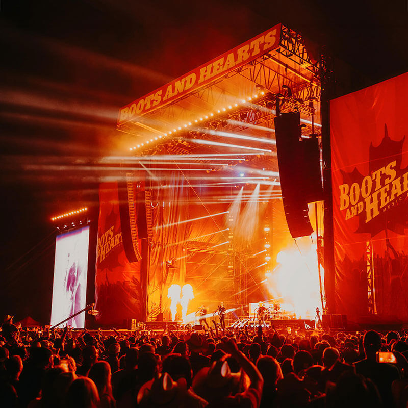 A large crowd watches a nighttime concert at the Boots and Hearts festival, with bright orange stage lights, fire effects, and a big screen displaying a performer. The stage is decorated with the festival’s logo.
