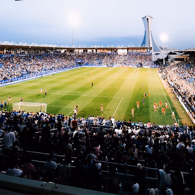 Vue depuis les tribunes lors d’un match du CF Montréal dans un stade de soccer professionnel, montrant un terrain vert occupé par des joueurs et une foule nombreuse installée dans les sièges bleus.