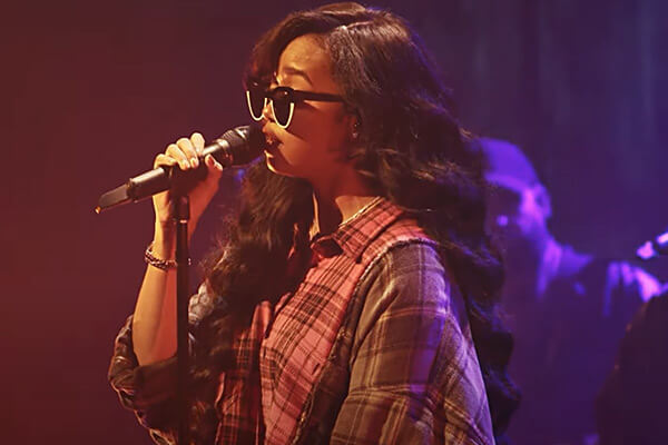 A woman with long wavy hair and sunglasses sings into a microphone on stage, wearing a plaid shirt as part of the Small Stage Series. The background is dark with purple lighting, capturing the intimate energy of live events.