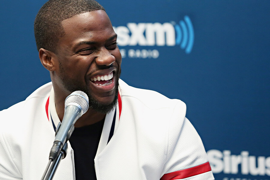 Kevin Hart is pictured in a white jacket laughing while speaking into a microphone, with a blue SiriusXM radio backdrop behind him.
