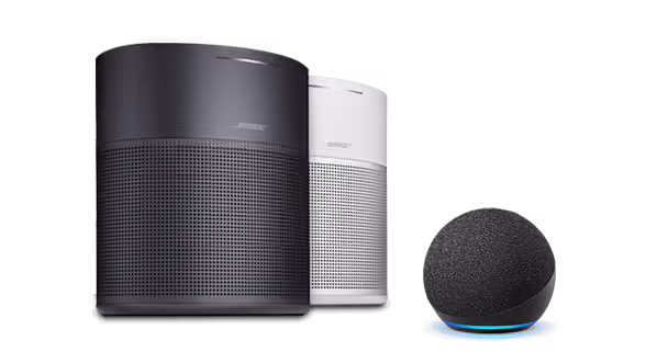Two Bose wireless speakers, one black and one white, stand side by side. Next to them is a small, spherical Amazon smart speaker with a glowing blue ring at its base—compatible with Amazon Alexa.