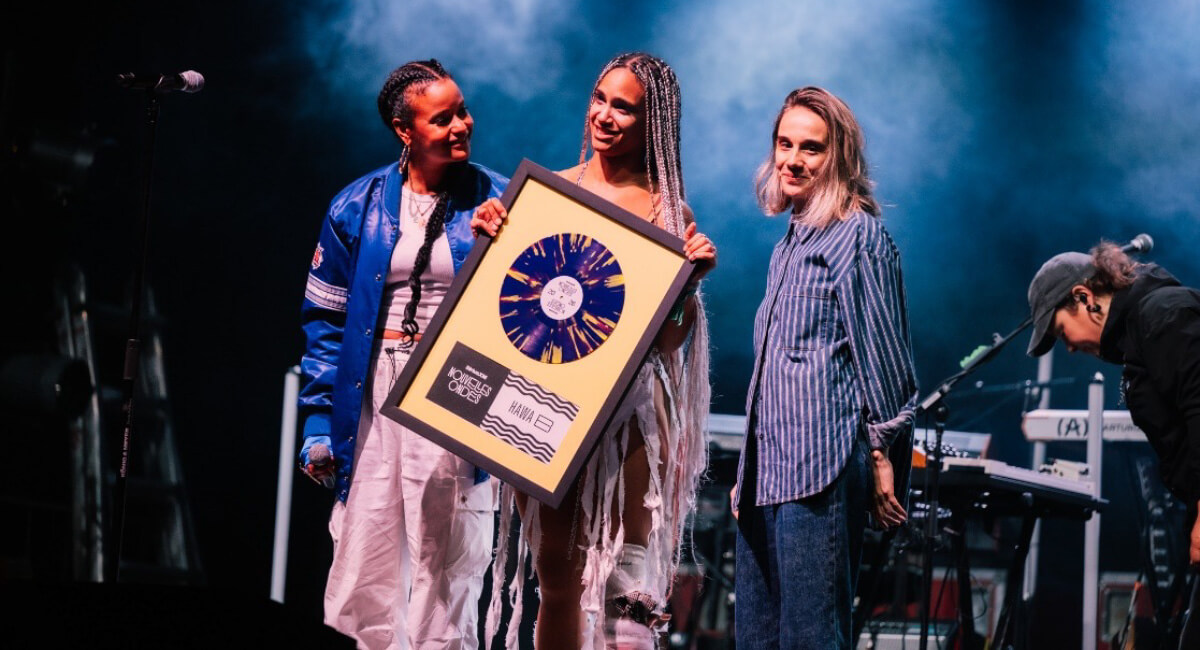 Marieme (animatrice), Hawa B (lauréate) et Matilde Bourgeois (responsable du développement de contenu canadien chez SiriusXM) sont toutes sur scène, souriantes et tenant une grande plaque de récompense, entourées de matériel musical.