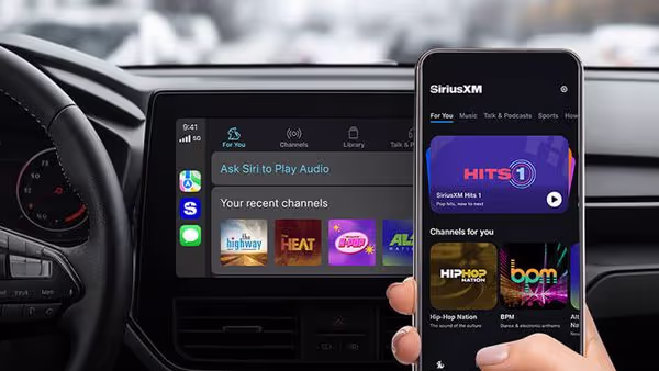 A person holds a smartphone displaying the SiriusXM app using Apple CarPlay in front of a car dashboard screen showing music apps and the prompt, Ask Siri to Play Audio.