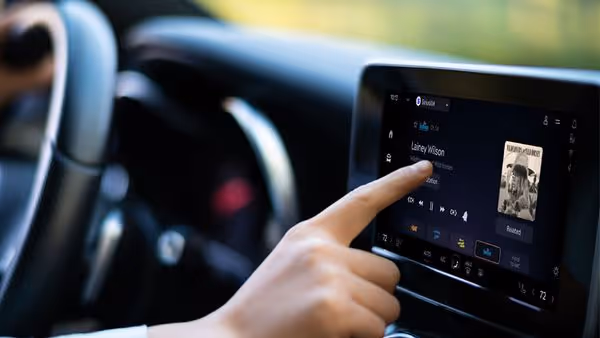 A person touches a car’s 360L radio touchscreen display, selecting a SiriusXM channel. The screen shows a song by Lainey Wilson with album art. The car interior and blurred scenery outside are visible.