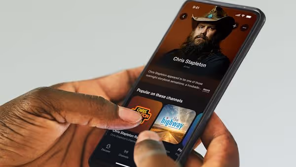 A person holds a smartphone displaying a the SiriusXM app featuring Chris Stapleton Radio, showing his photo and related channels like The Highway.
