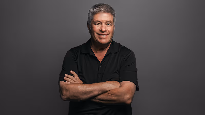 Gord Stellick is shown with short gray hair standing against a plain grey background, wearing a black polo shirt and smiling with his arms crossed. You can hear Gord Stellick on SiriusXM's Morning Skate podcast along with hosts Scott Laughlin and Tyler Madarasz.
