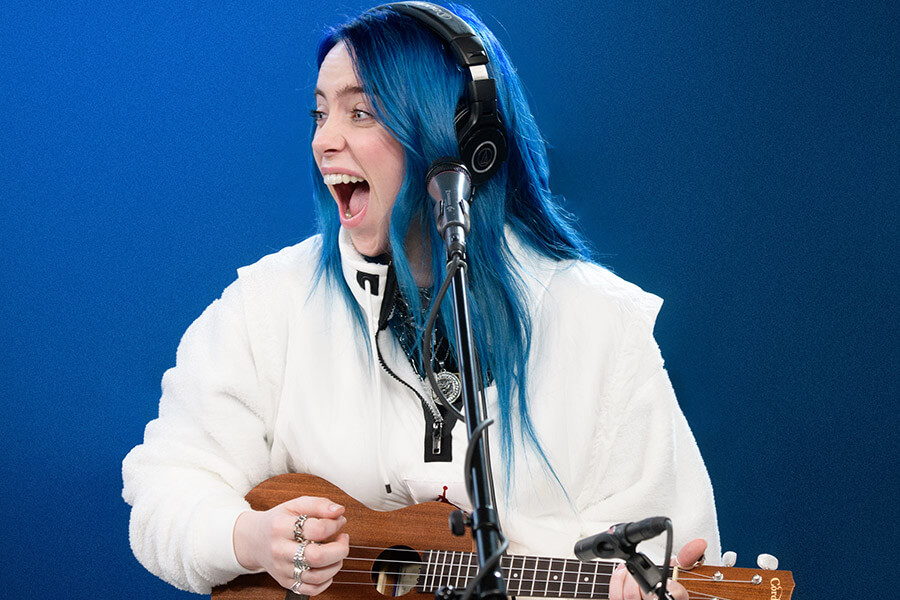 Billie Eilish is pictured with long blue hair wearing headphones and a white jacket smiles widely while playing a ukulele and standing in front of a microphone against a blue background.