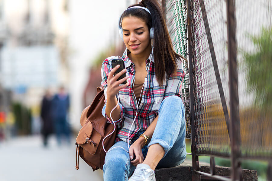Young woman with headphones sits outdoors, leaning against a fence, wearing a plaid shirt and jeans, smiling while looking at her phone. She has a brown backpack and is listening to music.